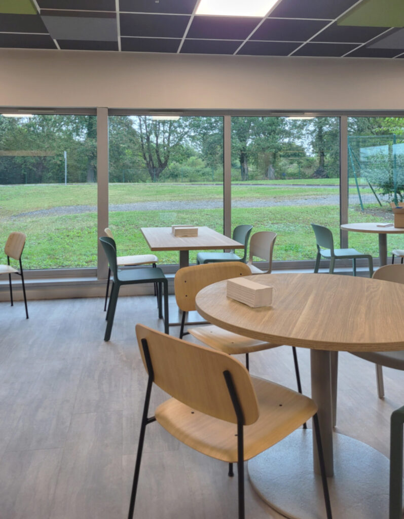Salle de pause d’entreprise avec tables et chaises près de grandes baies vitrées donnant sur un espace vert.