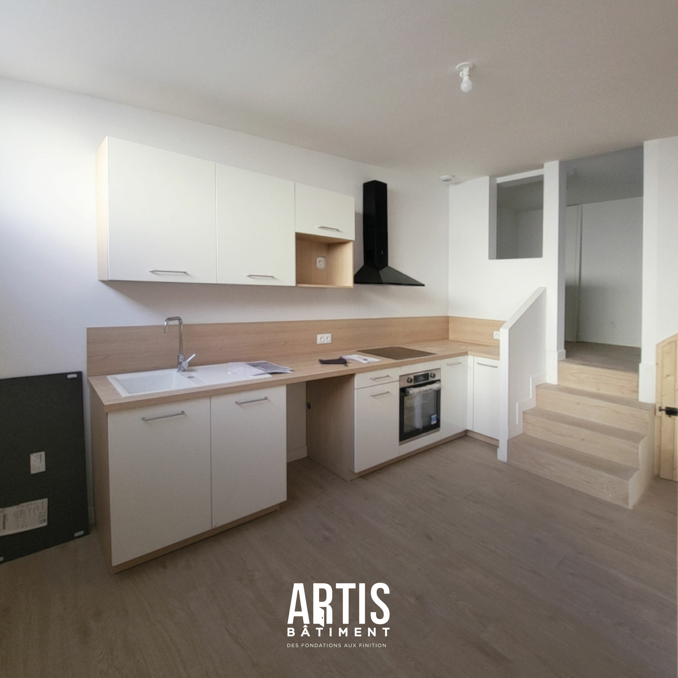 Cuisine ouverte rénovée avec mobilier blanc et bois, hotte noire et escalier intégré dans la pièce de vie.