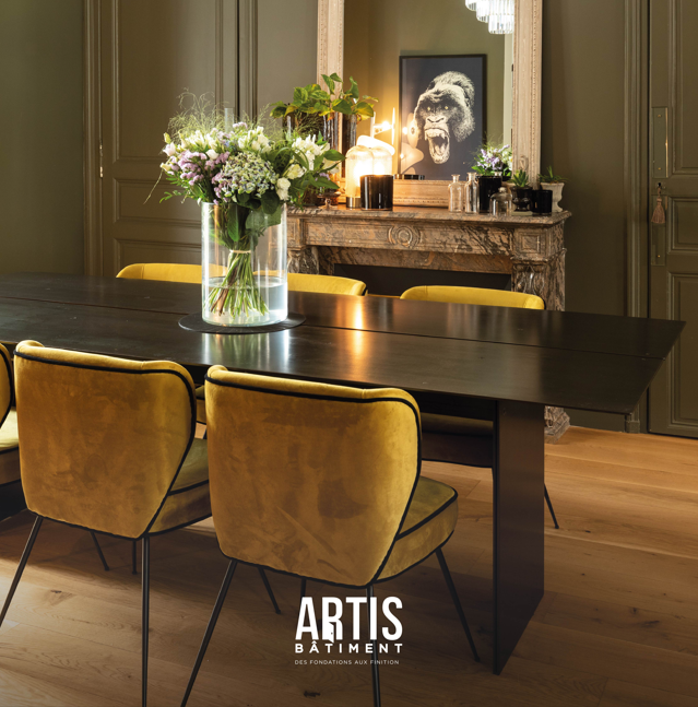 Salle à manger rénovée avec grande table noire, chaises velours jaunes et cheminée ancienne.