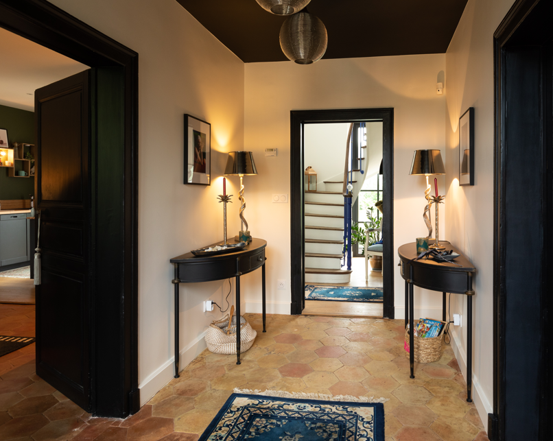Hall d’entrée d’une maison rénovée avec carrelage ancien, consoles noires et vue sur l’escalier.