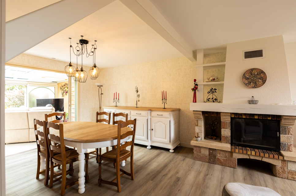 Salle à manger rénovée avec table en bois, cheminée en pierre et mobilier blanc.
